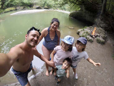 A imagem mostra uma família de cinco pessoas posando para uma foto na margem de um rio, com uma fogueira acesa ao fundo e árvores ao redor.
