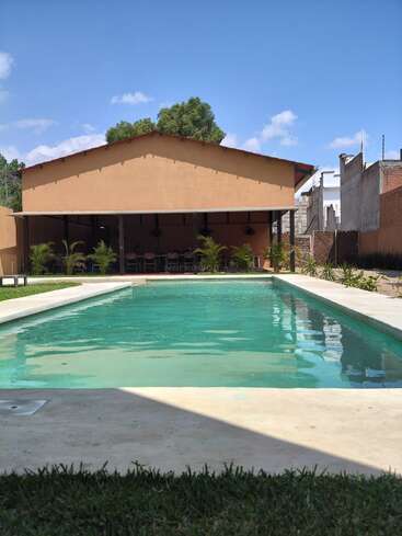 Una piscina rectangular de aguas cristalinas se encuentra frente a un edificio de color beige, rodeada de césped, plantas y sillas de exterior bajo un porche cubierto.