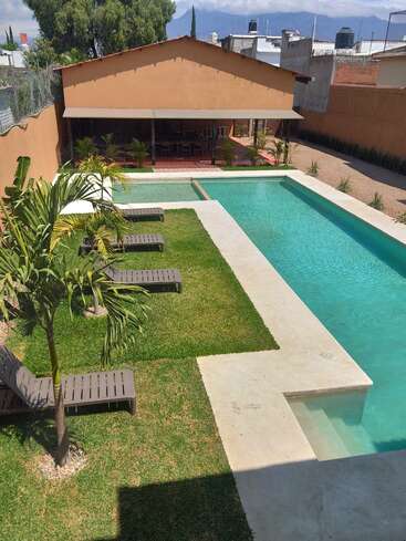 Un tranquilo jardín trasero cuenta con una piscina rectangular, césped verde, palmeras, varias tumbonas y una zona de patio a la sombra con mesas y sillas para relajarse.