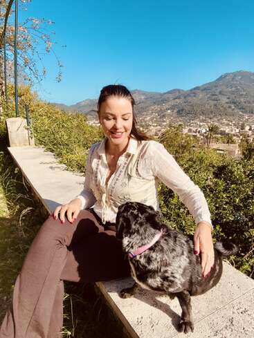Eine Frau sitzt draußen auf einer Steinbank, lächelt und streichelt einen schwarz-weißen Hund. Der blaue Himmel, das Grün und die Berge machen einen wunderschönen Hintergrund.