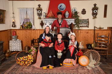 Eine fünfköpfige Familie in typisch niederländischer Tracht posiert in einem gemütlichen Vintage-Zimmer, das mit Tulpen, Käserädern, Holzschuhen und einer Ziehharmonika dekoriert ist.