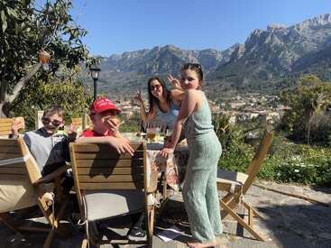 A group of people enjoy an outdoor meal together, smiling and posing by a table. Stunning mountains and a village provide a beautiful scenic background under sunny skies.