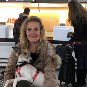 A smiling woman sits at an airport with a dog on her lap. People, luggage, and Swiss airline check-in counters are visible in the background.