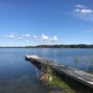 Ein hölzerner Steg ragt in einen ruhigen See unter einem strahlend blauen Himmel, mit vereinzelten weißen Wolken und entfernten grünen Bäumen am Horizont, eine friedliche Szene.