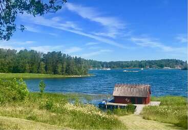 A serene lakeside scene features a red boathouse, vibrant greenery, blue sky with wispy clouds, calm water, distant sailboats, and a peaceful natural setting.
