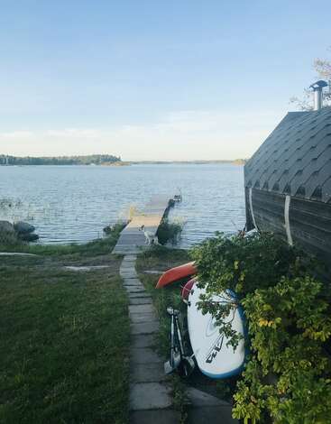 A serene lakeside view with a narrow dock, a small dog standing at the edge, a wooden hut, green grass, water sports equipment, and clear sky.