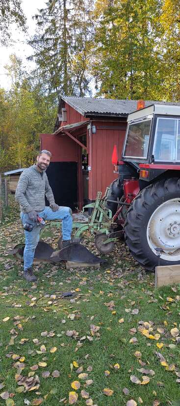 Ein lächelnder Mann steht draußen neben einem Traktor, hat ein Elektrowerkzeug in der Hand und einen Fuß auf einem landwirtschaftlichen Gerät. Herbstblätter liegen auf dem Gras, hinter ihm steht ein roter Schuppen.