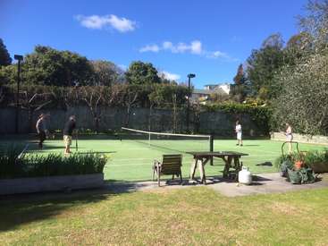 La imagen representa un sereno escenario al aire libre con una pista de tenis, en la que cuatro personas juegan, rodeadas de una exuberante vegetación y un cielo azul despejado.