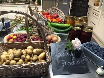La imagen muestra un mostrador de cocina con cestas de productos agrícolas, tarros de conservas y un cartel de pizarra que dice "Today's Produce" en tiza blanca.