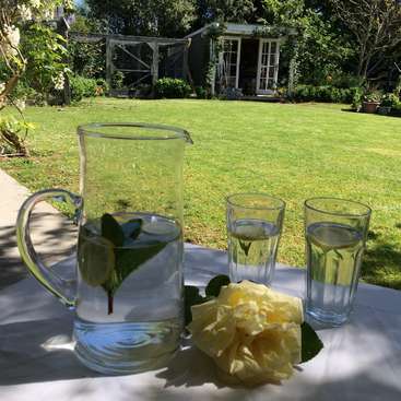 La imagen representa un entorno sereno al aire libre, con una jarra de agua con limón y menta, acompañada de dos vasos y una rosa blanca sobre una mesa, rodeada de un exuberante césped verde y una pequeña caseta de jardín.
