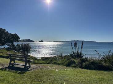 A wooden bench overlooks a sparkling, sunlit ocean. Blue sky, distant islands, green grass, and coastal plants create a peaceful, inviting seaside landscape under bright sunlight.