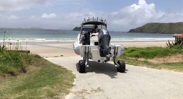 A boat with wheels is being driven down a ramp toward a sandy beach. The ocean and green hills are visible under a partly cloudy sky.
