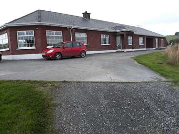 A imagem mostra uma casa de tijolos vermelhos de um andar com telhado cinza, com um carro vermelho estacionado na entrada da garagem, cercada por uma área gramada e um caminho de cascalho.