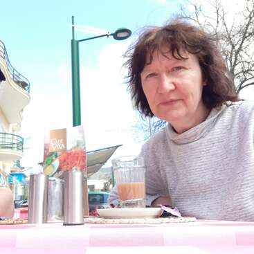The image shows a woman sitting at an outdoor table with a glass of orange liquid and a menu, set against a streetlight and buildings in the background.