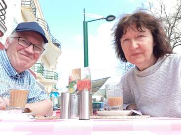 A imagem mostra um casal de idosos sentado em uma mesa com bebidas e comida, em frente a um poste de luz e prédios, em um dia ensolarado.