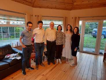 A imagem mostra um grupo de cinco pessoas em uma sala aconchegante com piso e paredes de madeira, cercada por janelas e uma porta, com um sofá e cadeiras ao fundo.