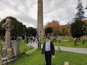 A imagem mostra um homem em pé em um cemitério, cercado por lápides e uma alta torre de pedra, com um grupo de pessoas caminhando ao fundo.
