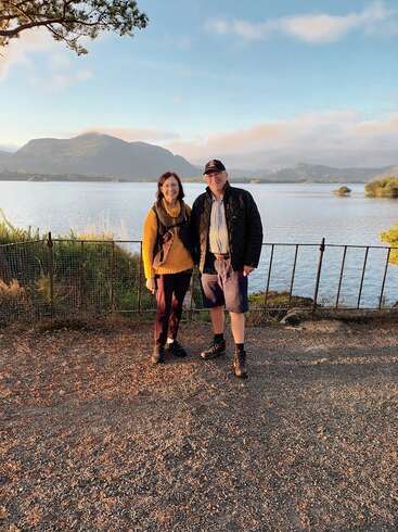A imagem retrata uma cena serena de um homem e uma mulher em pé em frente a um lago, com montanhas visíveis ao fundo, em um céu azul com nuvens.