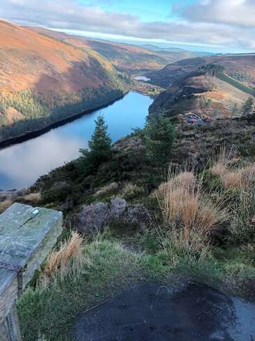 A imagem retrata uma paisagem serena com um lago aninhado entre colinas onduladas, com um caminho sinuoso e vegetação exuberante, em um céu nublado.