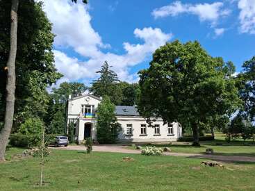 Ein weißes Haus steht umgeben von hohen grünen Bäumen und einem großen grasbewachsenen Hof. Ein silbernes Auto ist auf der Auffahrt geparkt. Der Himmel ist strahlend blau.