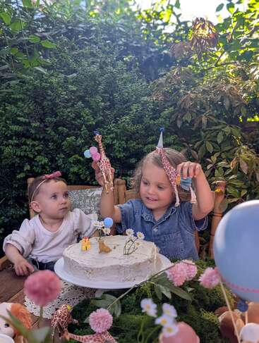 Dos niños pequeños celebran al aire libre con una tarta decorada con flores y animalitos de juguete. Uno de ellos lleva un gorro de fiesta y sostiene con alegría unas figuritas de jirafas en alto.