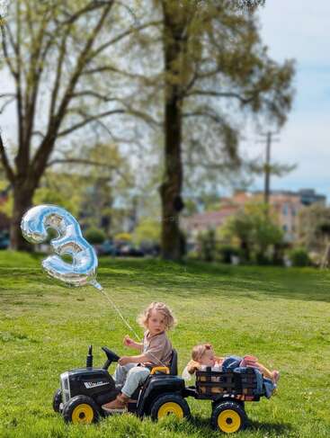 Dos niños pequeños juegan al aire libre en un césped. Uno conduce un tractor de juguete con un globo plateado con el número «3», mientras que el otro descansa en el remolque acoplado.