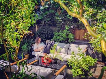Una mujer descansa en un acogedor sofá al aire libre, rodeada de exuberantes plantas y árboles, leyendo, con una taza de café sobre una mesita cercana.