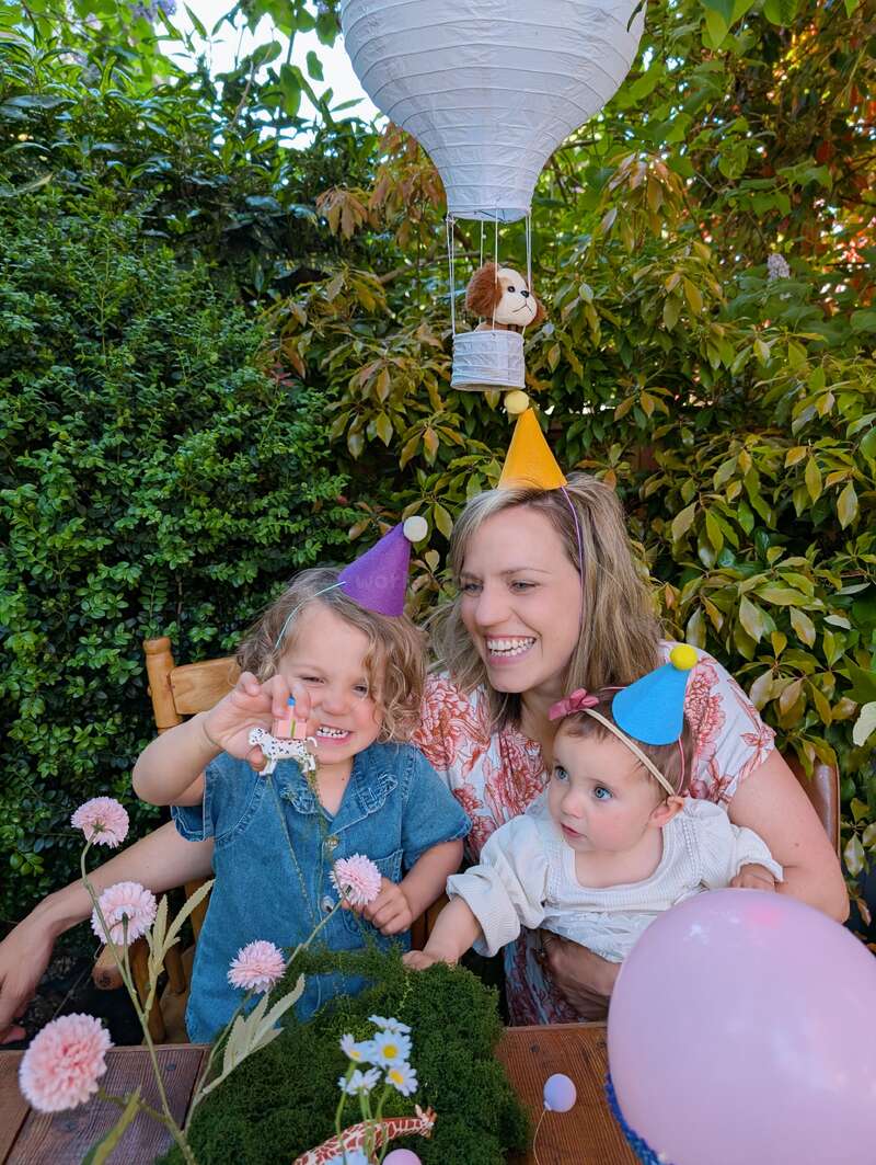 Una madre radiante y sus dos hijos pequeños celebran al aire libre, con coloridos gorros de fiesta. Están rodeados de vegetación, flores, un globo y una divertida decoración con globos aerostáticos.