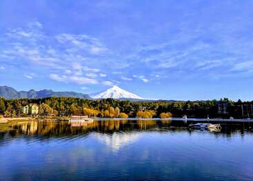 In einem ruhigen See spiegeln sich bunte Bäume und Häuser. Im Hintergrund erhebt sich majestätisch ein schneebedeckter Vulkan unter einem strahlend blauen, wolkenverhangenen Himmel.