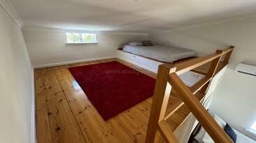 Esta imagem mostra um quarto loft aconchegante com teto baixo, pisos de madeira, um tapete vermelho, um colchão em um estrado e uma pequena janela que fornece luz natural.