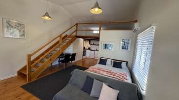 Esta imagem mostra um apartamento estúdio aconchegante com um loft de madeira, cama de casal, sofá cinza, mesa de jantar, cozinha moderna, obras de arte e janelas grandes com persianas.