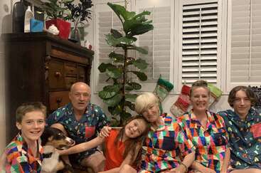 Uma família usando pijamas coloridos combinando está sentada em uma sala aconchegante, sorrindo, com seu cachorro, meias e uma planta decorada, comemorando um feriado festivo.