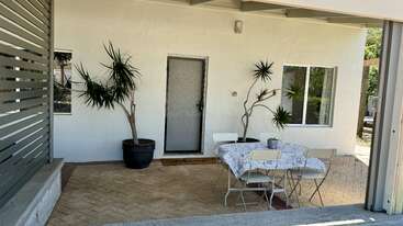 Um pátio branco tem dois vasos de plantas, uma pequena mesa coberta com um pano, quatro cadeiras, um tapete e uma porta com um painel de vidro fosco.