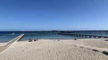 Una soleada playa de suave arena blanca desemboca en un tranquilo mar azul. Varios muelles de madera se adentran en el agua. La gente se relaja y se baña. Ambiente tranquilo.