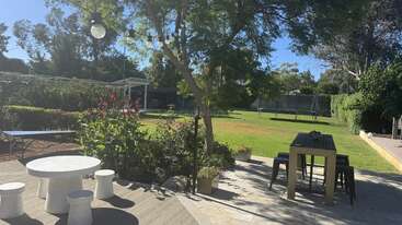 Un patio soleado con césped verde, árboles, plantas con flores, luces de cuerda para exteriores, una mesa blanca redonda con taburetes, una mesa de comedor de madera y una cama elástica. Ambiente tranquilo.