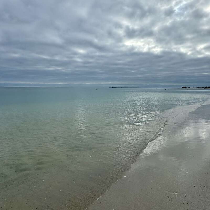A água calma do mar toca suavemente a costa arenosa sob um céu nublado. A luz suave reflete nas ondas azul-esverdeadas pálidas. O horizonte parece pacífico e distante.