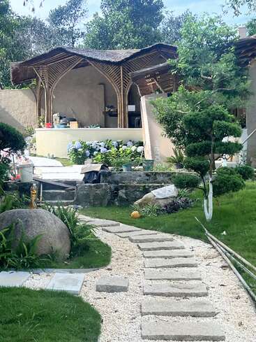 Un chemin de pierre incurvé traverse un jardin verdoyant avec des buissons taillés, des rochers et des fleurs, vers un pavillon en bambou et des matériaux de construction partiellement visibles.
