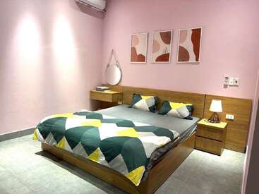 Une chambre à coucher confortable avec des murs rose pastel, des meubles en bois, une literie géométrique, un miroir rond, une lampe de chevet et des œuvres d'art abstraites, créant une atmosphère moderne et accueillante.