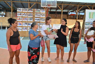 A group of women stands together on a basketball court, smiling as one receives a certificate. Photo boards and a CAEMBA banner are displayed behind them.