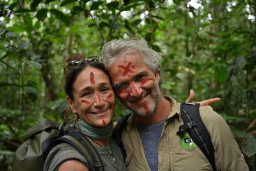 A smiling couple stands close together in a lush, green forest. Both have red face paint markings and wear backpacks, suggesting an adventurous outdoor journey.