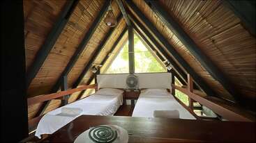 This cozy attic room features two single beds with white linens, wooden and bamboo walls, triangular ceiling, large window, a fan, and soft natural lighting.