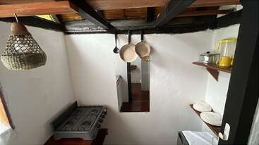 This cozy kitchen features white walls, wooden ceiling beams, a small gas stove, hanging utensils, open shelves with dishes, a yellow kettle, and a woven pendant lamp.