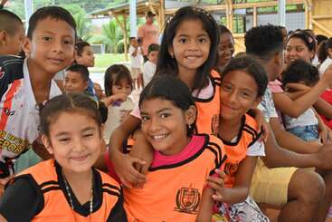 A group of happy children pose together, smiling warmly. They wear orange sports jerseys and are surrounded by other kids, creating a joyful and friendly atmosphere.