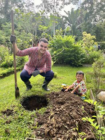 Um homem e uma criança estão jardinando juntos, sorrindo alegremente. O homem segura uma pá; a criança brinca com um caminhão de brinquedo ao lado de um buraco. Uma vegetação exuberante os rodeia.