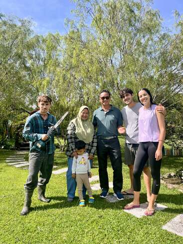 Seis pessoas estão juntas na grama verde com árvores atrás delas. Uma delas segura uma ferramenta elétrica. O grupo sorri, aproveitando um dia ensolarado ao ar livre.