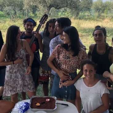 Eine Gruppe von Freunden versammelt sich im Freien, lächelt und plaudert, während einer Gitarre spielt. Auf dem Tisch steht ein Kuchen, der auf eine fröhliche Feier oder einen besonderen Anlass hindeutet.
