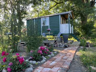 Das Bild zeigt eine kleine, grün-weiße Holzhütte mit einer Terrasse und einem gemauerten Weg, der zu ihr führt, umgeben von Bäumen und Blumen.