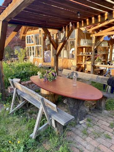 Das Bild zeigt eine ruhige Umgebung im Freien mit einem Holztisch mit einem Baumstammsockel, umgeben von Bänken und einer rustikalen Holzkonstruktion im Hintergrund.