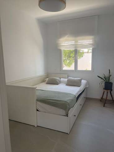 A bright, minimalist bedroom features a white daybed with storage drawers, green blanket, two pillows, a sunny window, white blinds, potted plant, and light fixture.