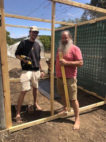 Dois homens estão descalços e sorridentes enquanto trabalham em uma estrutura de madeira ao ar livre. Um segura uma furadeira elétrica e o outro um nível de bolha de ar. Dia ensolarado, com árvores verdes atrás.
